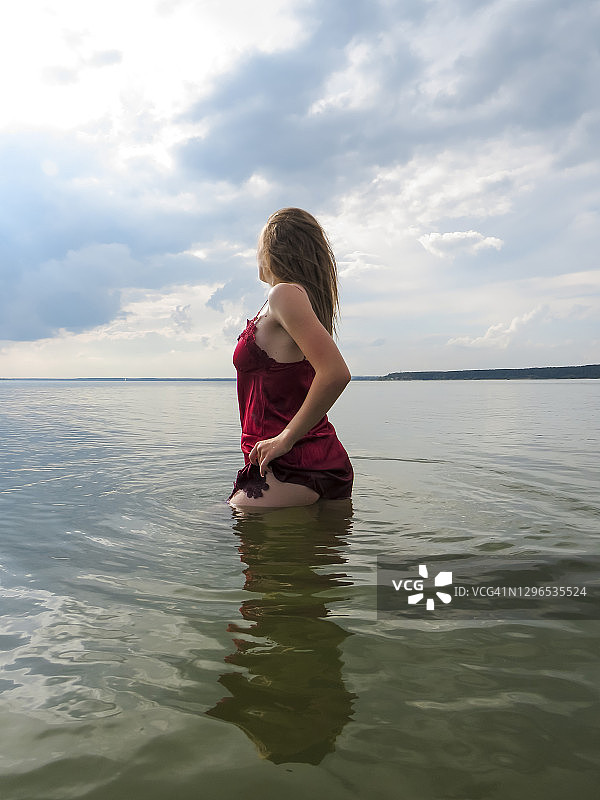 迷人的年轻金发女人松散的头发和性感苗条的身体在一个炎热的夏天，在一个浅滩上的水湿红色丝绸连衣裙图片素材