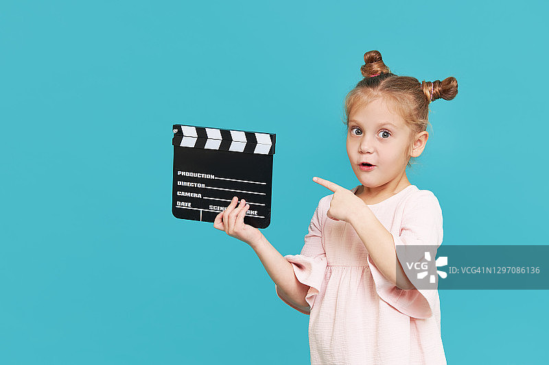 滑稽微笑的小女孩拿着电影制作板孤立在蓝色的背景。小剪匠，表演训练。有趣的脸。为文本复制空间。“t图片素材