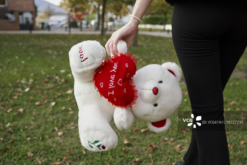 一个女人走着，手里拿着一个白色的泰迪熊，中间有一颗红心，上面写着“我爱你”。情人节的概念图片素材