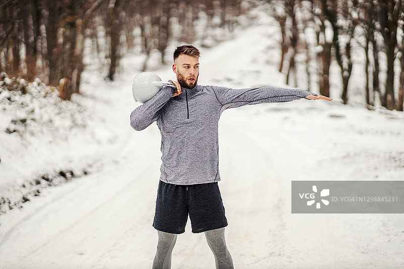 健硕的运动员站在林间积雪的小路上举壶铃。冬季健身，健康生活，健美健身图片素材