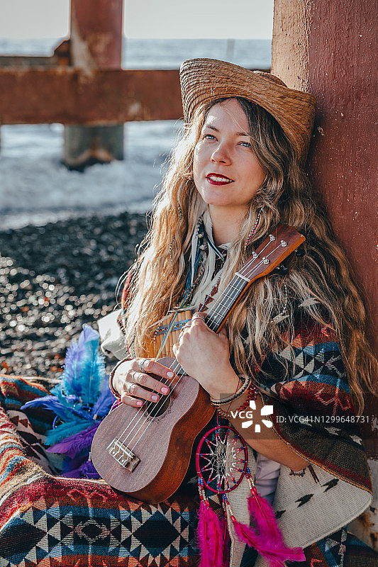 一个年轻的嬉皮女人坐在码头附近的海滩上弹奏四弦琴。图片素材