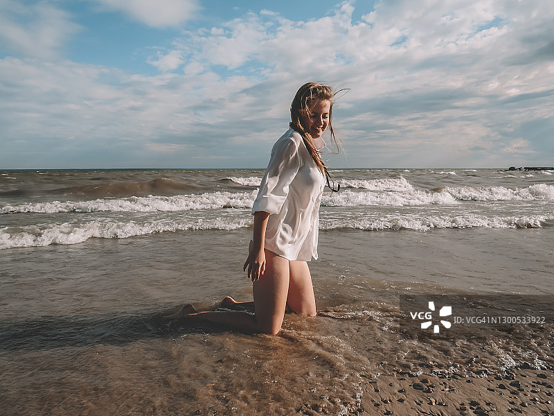 一个美丽的年轻女子与风吹头发在白色丝绸衬衫和比基尼在潮湿的沙滩上的海浪和暴风雨的天空的背景图片素材