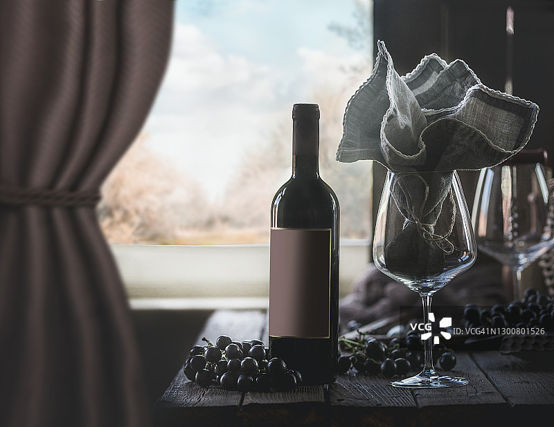 有空白标签的红酒瓶子，用亚麻餐巾和葡萄装饰的酒杯放在有窗帘和乡村景色的乡村木桌上图片素材