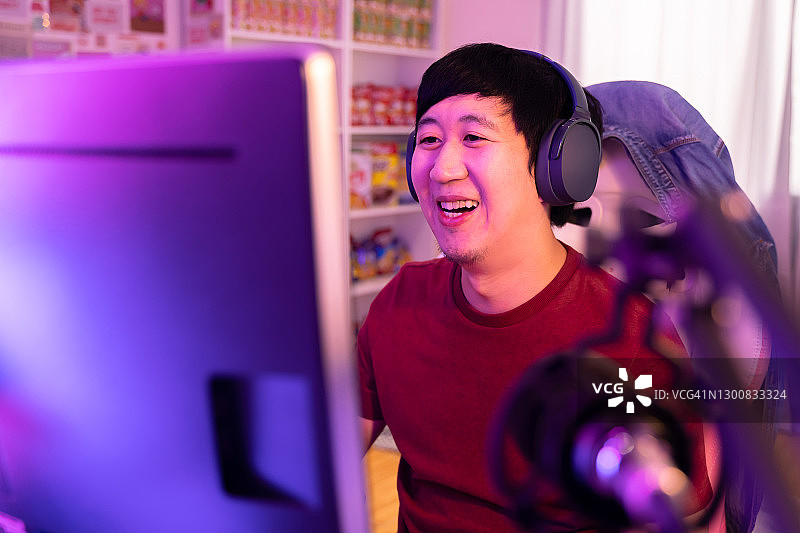兴奋和微笑的男性玩家戴着耳机麦克风玩在线视频游戏。年轻的亚洲人在家里用个人电脑与玩家和观众交谈图片素材