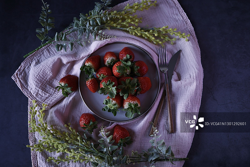 在深蓝色的石头地板上，灰色的盘子里放着新鲜的草莓，盘子下面是粉红色的镶边布餐巾，上面装饰着两朵人造的黄色野花，草莓散落在周围，盘子旁边放着一个刀叉图片素材