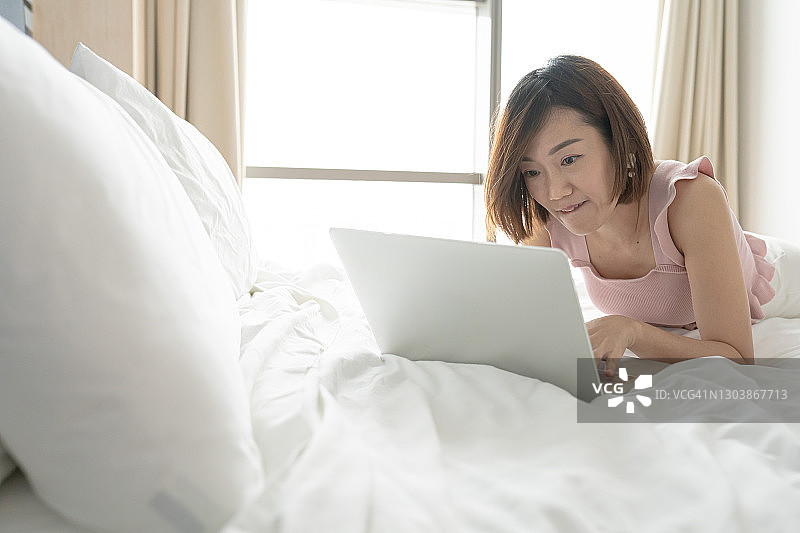 亚洲华人成熟的女人躺在床上使用笔记本电脑。放松图片素材