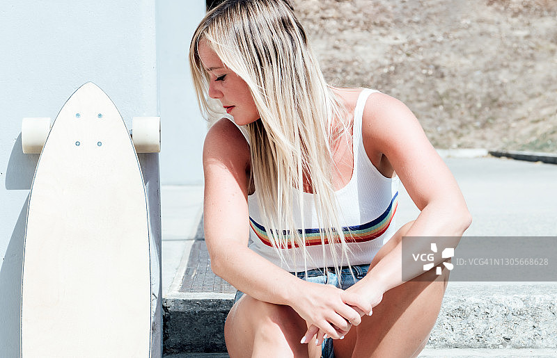 穿着短裤的金发女人坐在梯子上看着她旁边的长板滑板。图片素材