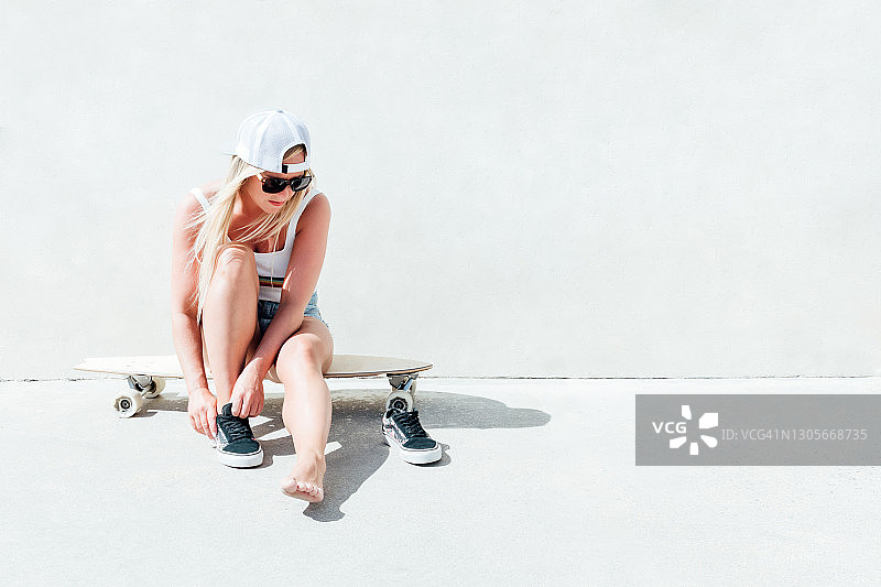 金发女人戴着太阳镜和灰色的帽子，坐在她的长滑板鞋上面。图片素材