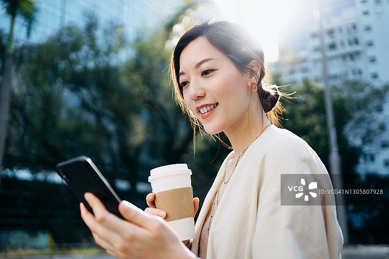 在清新的早晨，自信的年轻亚洲女商人一边走在城市的金融区，一边看电子邮件、喝咖啡图片素材