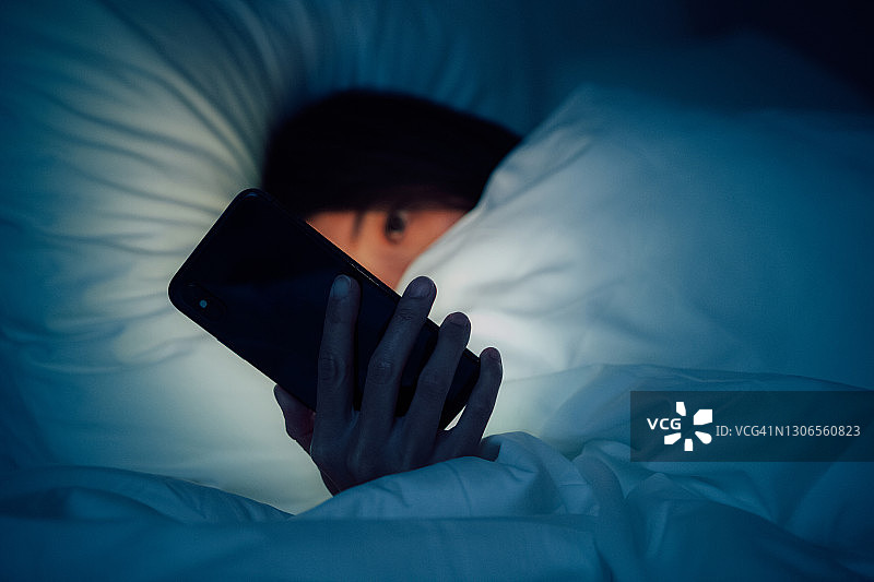 一个女人躲在毯子下面，深夜躺在床上玩智能手机图片素材