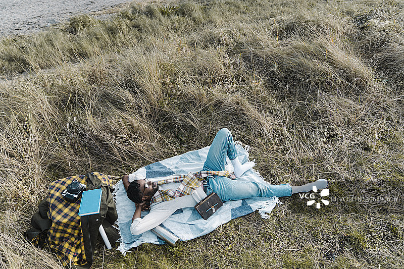 一名男子双手放在脑后，躺在沙滩上干燥的植物中间的毯子上休息图片素材