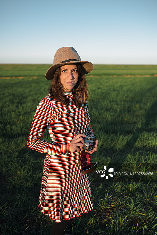 正面的一个年轻的白人妇女与她的老胶片相机走过一个谷物田，而拍照。图片素材