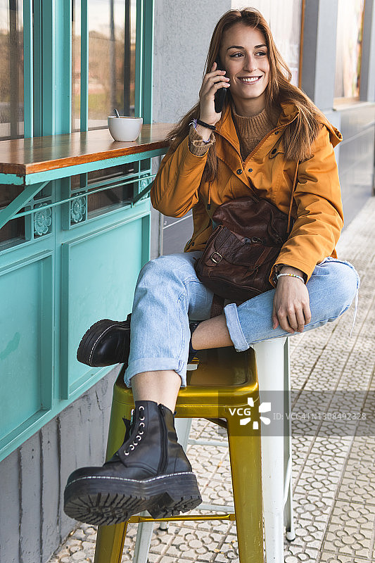 一个微笑的女人坐在路边咖啡馆的凳子上用手机聊天图片素材