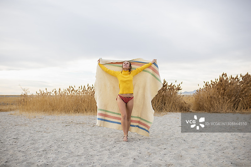 年轻女子拿着毯子，举起双臂走在海滩上图片素材