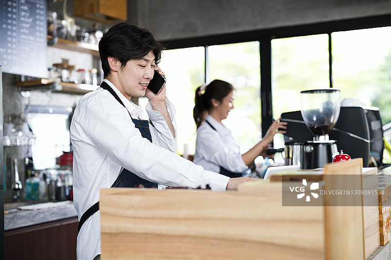 微笑的亚洲男性老板正在一间咖啡店里与顾客用手机点菜。餐饮点菜系统，客户服务头脑和小企业主。图片素材