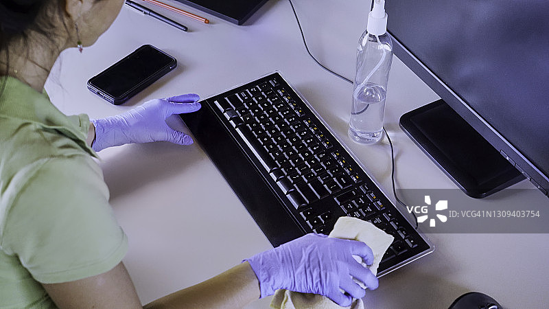 一名西班牙裔年轻女子在电脑键盘上喷洒并外用酒精消毒COVID-19图片素材