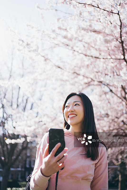 亚洲年轻开朗的女子，手拿智能手机，仰望天空，站在公园里盛开的樱桃树前图片素材