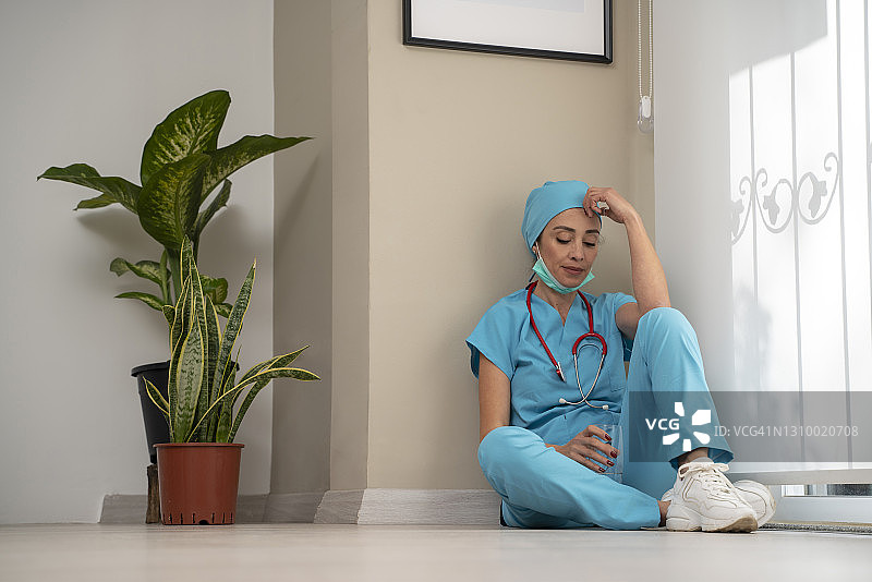 过度劳累的女医务人员戴着口罩、蓝色制服和手套坐在医院的地板上图片素材