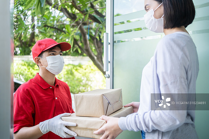 亚洲的邮递员、快递员戴着口罩把小箱子送到顾客家门口。男子戴口罩预防新型冠状病毒、冠状病毒情感爆发。送货上门的购物理念。图片素材