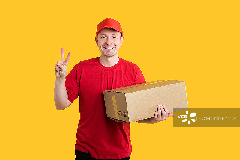 从网上商店送货上门。快乐的快递员在红色制服拿着一个棉花盒子，显示世界的标志在黄色孤立的背景在演播室。本空间图片素材
