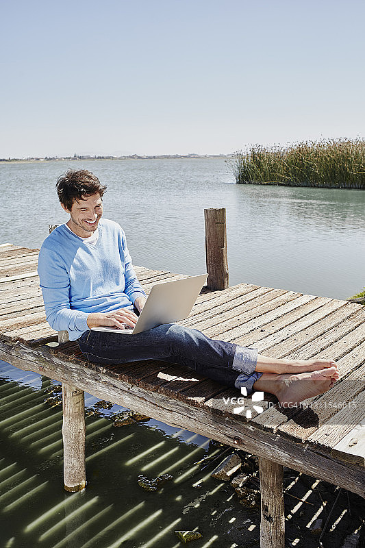 在阳光明媚的日子里，一名微笑的男子坐在码头上使用笔记本电脑图片素材