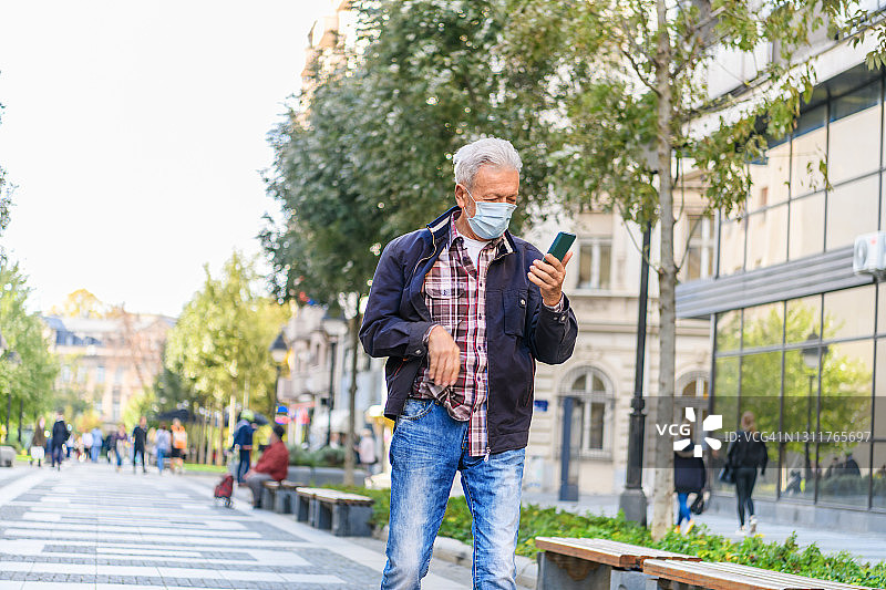 一位戴着口罩的老人走在城市的街道上并使用他的智能手机。图片素材