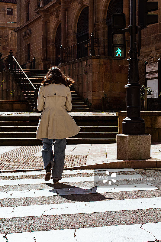 年轻女子穿雨衣穿过人行道/人行横道与绿色semaro在城市的后视图图片素材