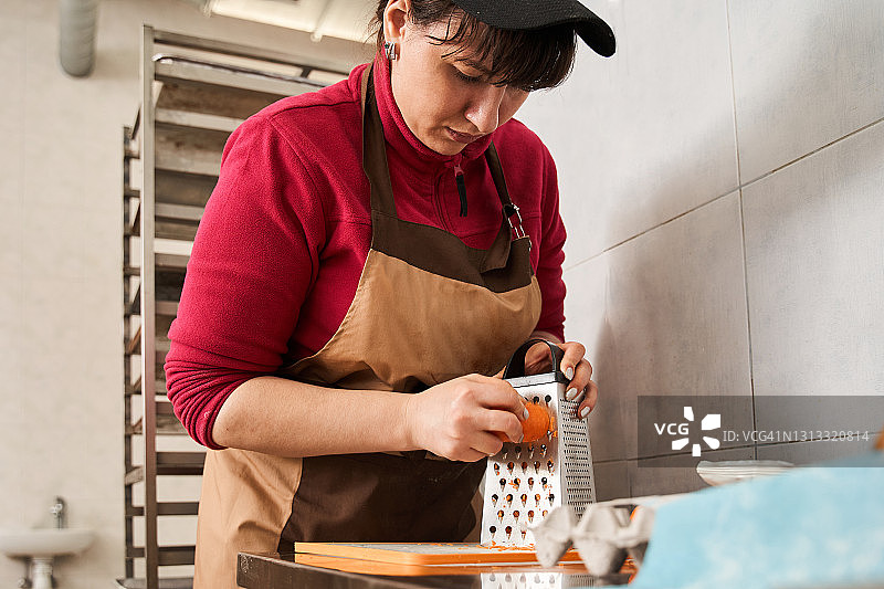 一位穿着围裙的女面包师在刨丝器上擦橘子皮图片素材