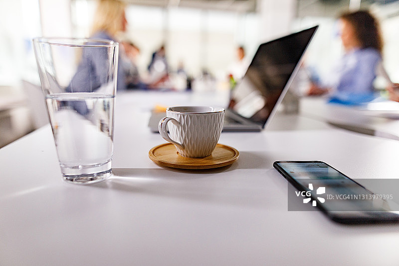 咖啡杯，一杯水和手机放在办公室的会议桌上。图片素材