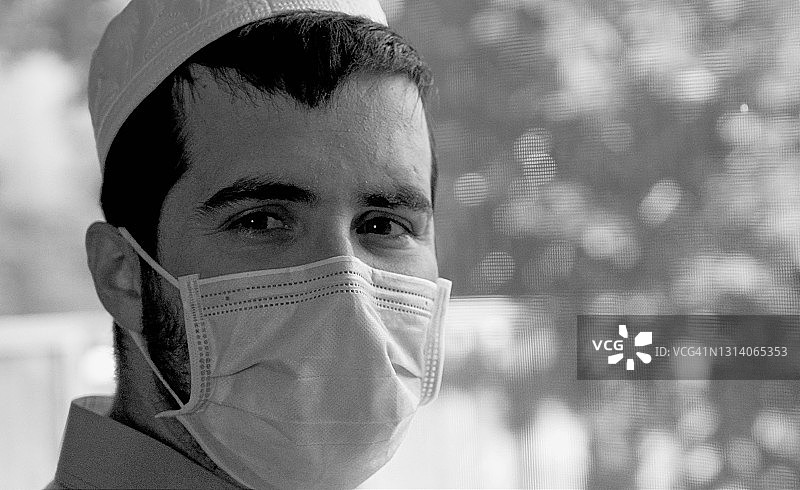 一名沉思的留着胡子的中东青年戴着防护面罩看着窗户附近的摄像头图片素材