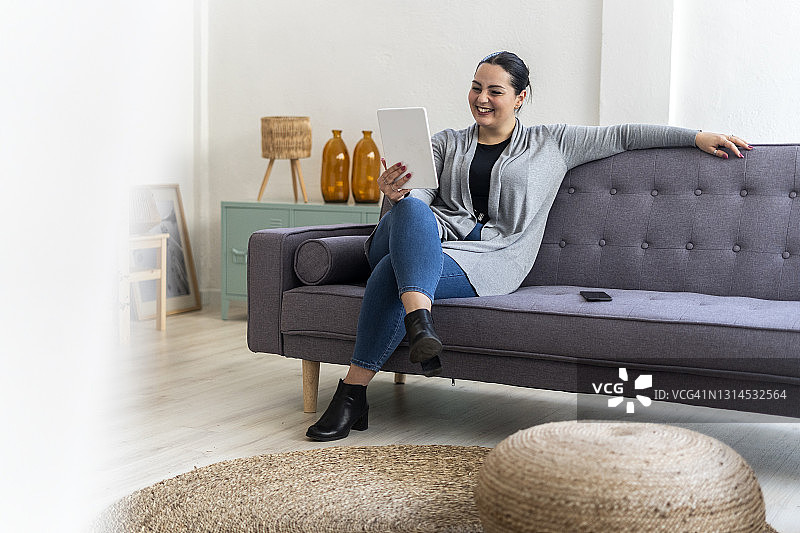 微笑的女人坐在客厅的沙发上使用数码平板电脑图片素材