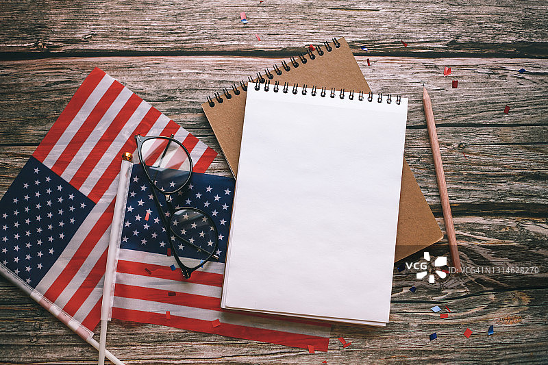 美国国旗、五彩纸屑、有拷贝空间的便笺本、眼镜和木制背景的铅笔。7月4日，独立日。图片素材