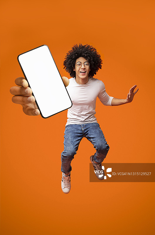移动应用广告。兴奋的青少年黑人男子显示智能手机空屏幕，跳跃在橙色背景图片素材