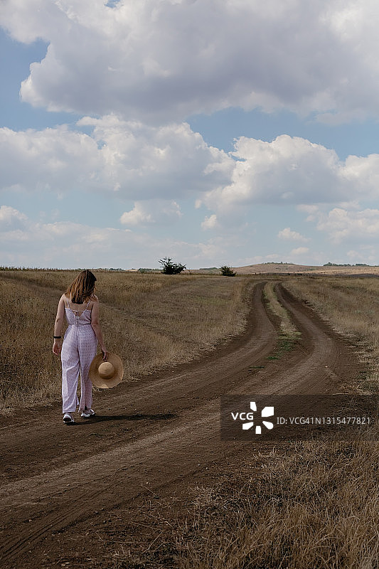 一个女孩沿着一条路走在一片干燥的草地上。后面的观点。图片素材