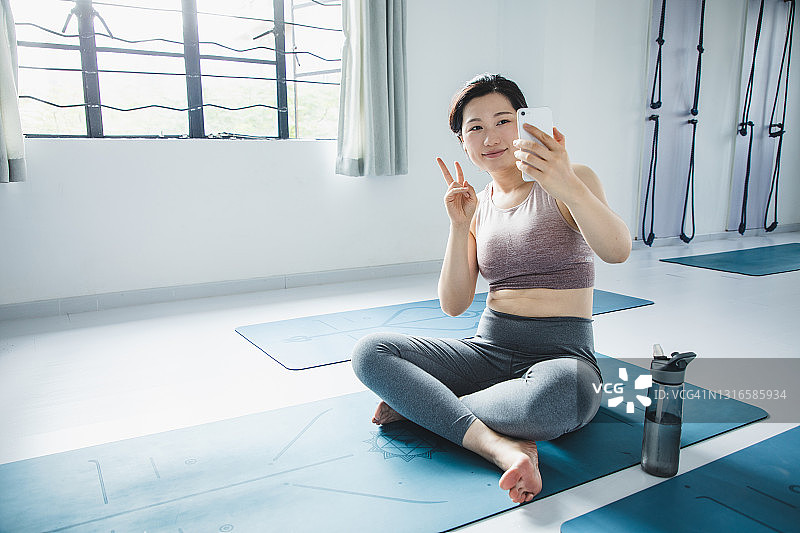 一名亚洲女子坐在瑜伽垫上，练完瑜伽后用手机自拍图片素材