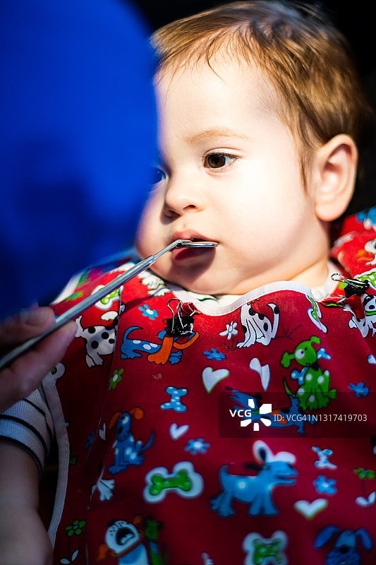 一个两岁以下的孩子在一个穿着红色长袍的牙医的办公室里，当孩子看着牙医的时候，牙医正试图用一面小镜子打开孩子的嘴。选择性方式。图片素材