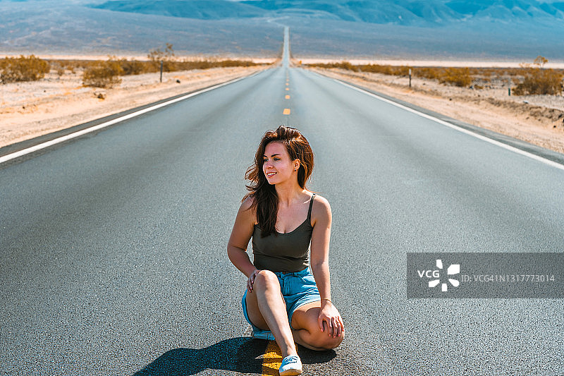 美丽的年轻女子与长头发走在一个风景如画的空道路在死亡谷俯瞰山脉，美国图片素材