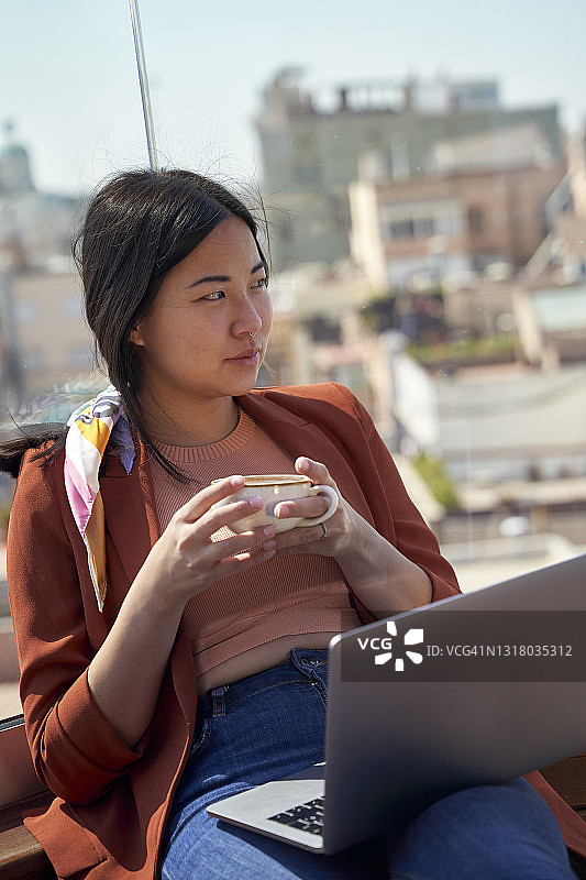 体贴的女人坐在屋顶露台上，拿着笔记本电脑和咖啡杯图片素材