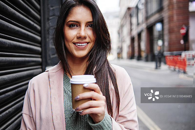 微笑的年轻女子拿着一次性咖啡杯站在城市的百叶窗旁图片素材
