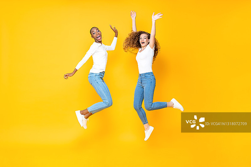 两个跨种族的女人朋友自由跳跃在孤立的工作室黄色背景图片素材