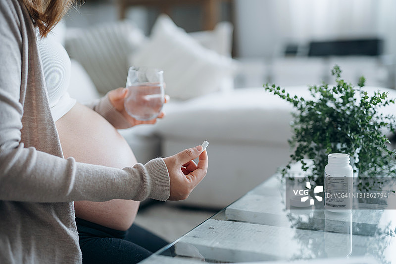 年轻的亚洲孕妇坐在家里客厅的沙发上，手里拿着药，手里拿着一杯水图片素材