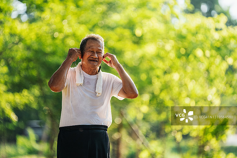 亚洲老年人的肖像穿着健身服在公园里跑步。一个微笑的男人的特写，一边跑步一边用耳机听音乐。图片素材