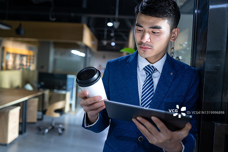 亚洲年轻男性经理在办公室拿着平板电脑，手里拿着咖啡杯图片素材