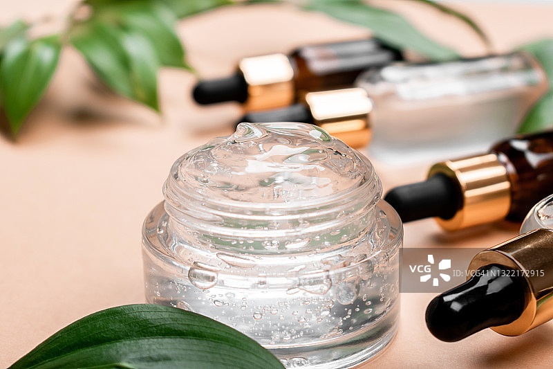 一瓶精致透明的保湿面凝胶，面霜和瓶面血清在柔和的米色背景与绿叶。护肤和化妆用的化妆品。图片素材