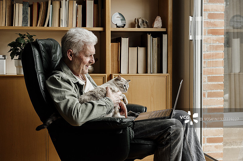 一位老人坐在家里的椅子上，拿着笔记本电脑，抱着一只猫图片素材