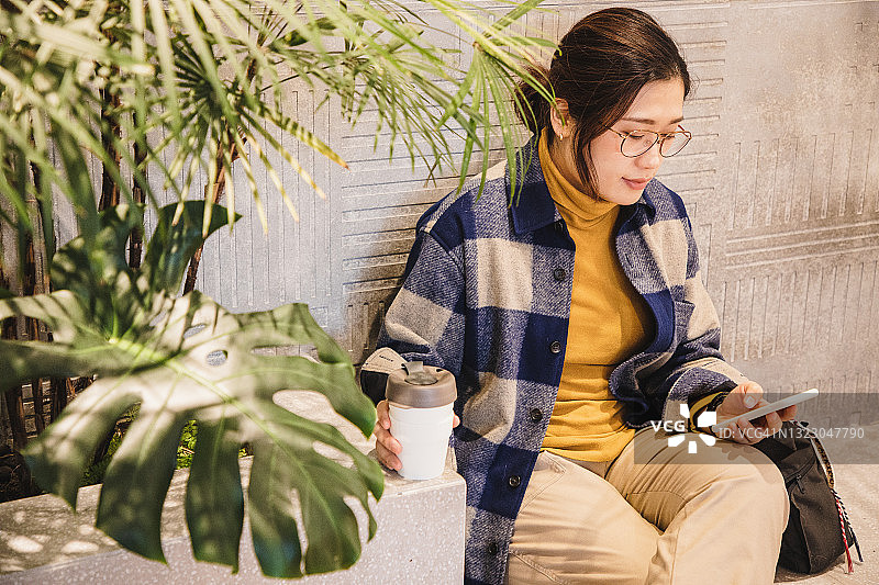 亚洲女性坐着使用电话，用可重复使用的咖啡杯喝咖啡图片素材