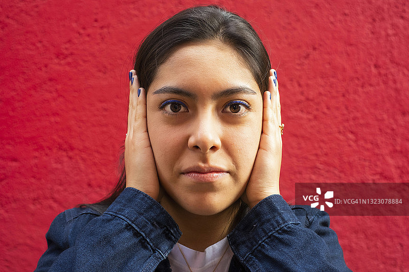 年轻的拉丁裔妇女看着镜头，遮住她的耳朵，听不到邪恶，红色背景图片素材