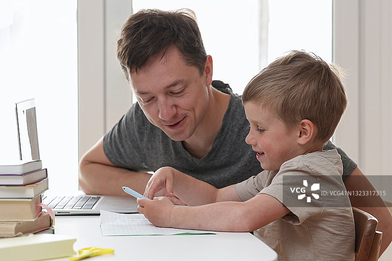 在新冠肺炎疫情隔离期间，父亲帮助孩子在电脑上做作业。自我孤立。年轻的爸爸和儿子在家里用平板电脑学习。图片素材