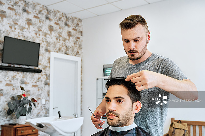 在一家理发店里，一位年轻的理发师正在用剪刀给一个年轻人理发。图片素材