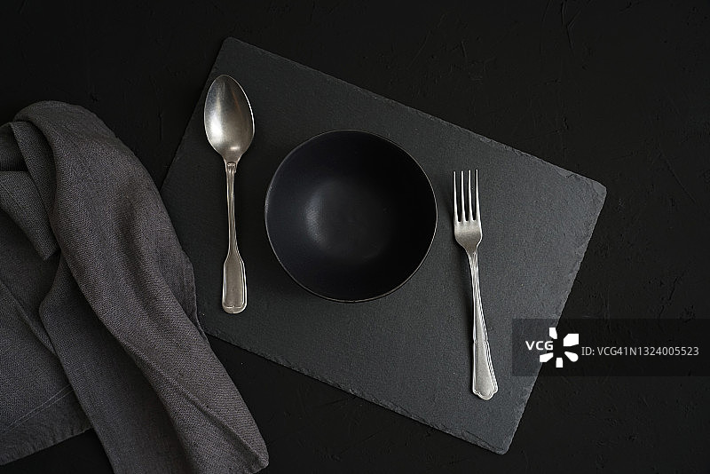 空的黑色陶瓷碗，复古的餐具和灰色亚麻织物在深色石板的背景图片素材